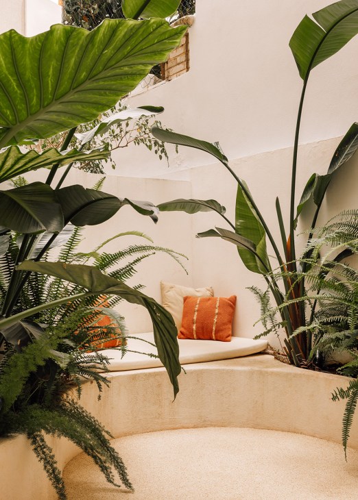 A minimalist courtyard with tropical plants, white gravel, and a cream-colored seating area with an orange cushion.