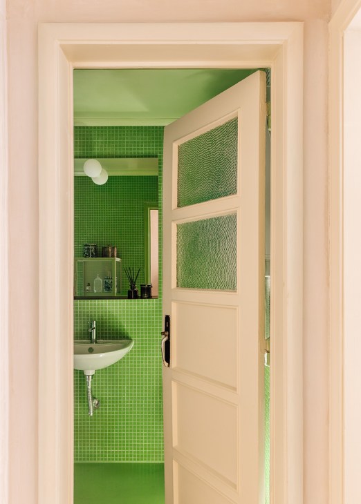 Bright green tiled bathroom with cream-colored door, wall-mounted sink, and shelving with bath products and decorative items.