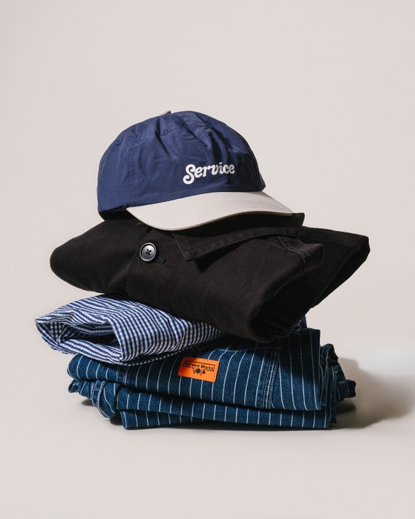 Service Works navy and cream cap with striped shirt, black jacket and pinstriped workwear stacked neatly.