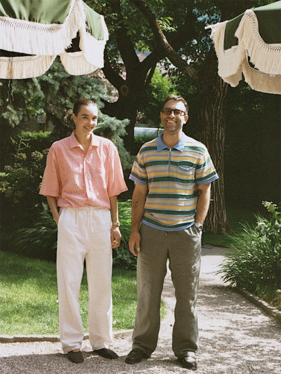 Two people stand in a garden under hanging macramé decorations; woman in pink shirt and cream pants, man in striped shirt and tan pants.