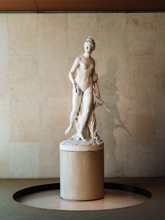 White marble sculpture of Diana by Jean-Antoine Houdon displayed on a cylindrical pedestal against a stone wall.