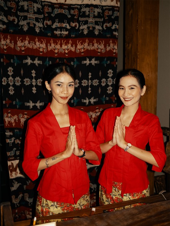 Two women in red traditional kebayas with golden patterned sarongs greet guests with prayer hands in front of ornate batik wall hangings.