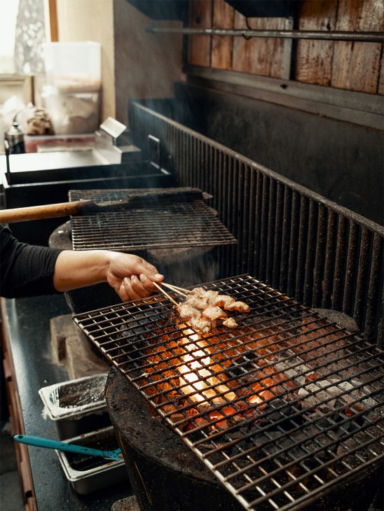 A chef's hand uses tongs to grill skewered meat over charcoal on a metal grate.