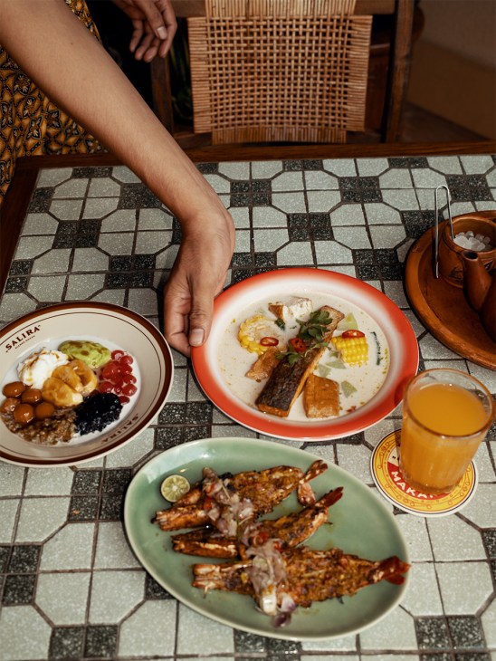 Overhead view of Indonesian dishes at Salira restaurant including grilled fish, soup, and rice with beverages.
