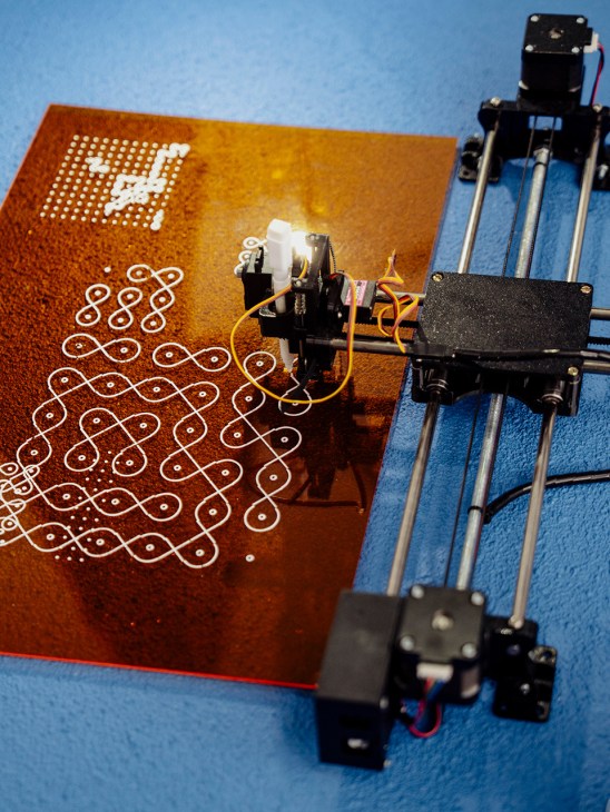 A 3D printer with orange and blue coloring printing a patterned design on a copper-colored substrate.