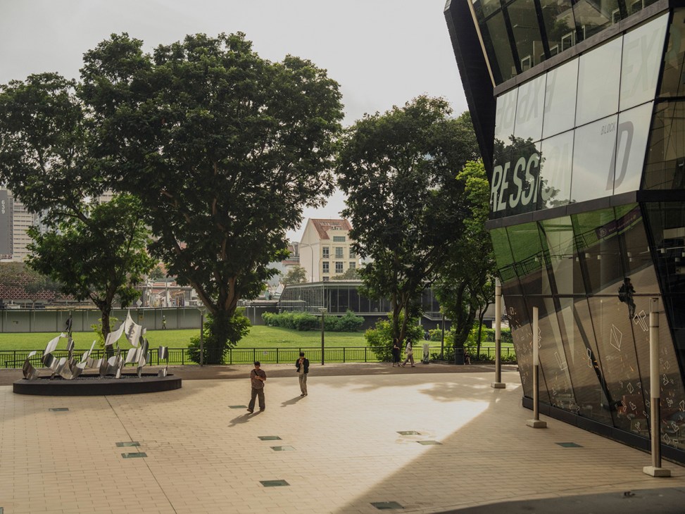 Outdoor plaza at Lasalle College of the Arts with paved courtyard, large trees, and modern building with glass facade.