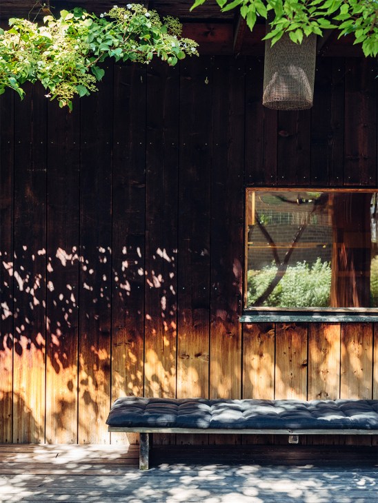 Dark wooden exterior wall with dappled sunlight shadows and a blue cushioned bench beneath a window overlooking a garden.