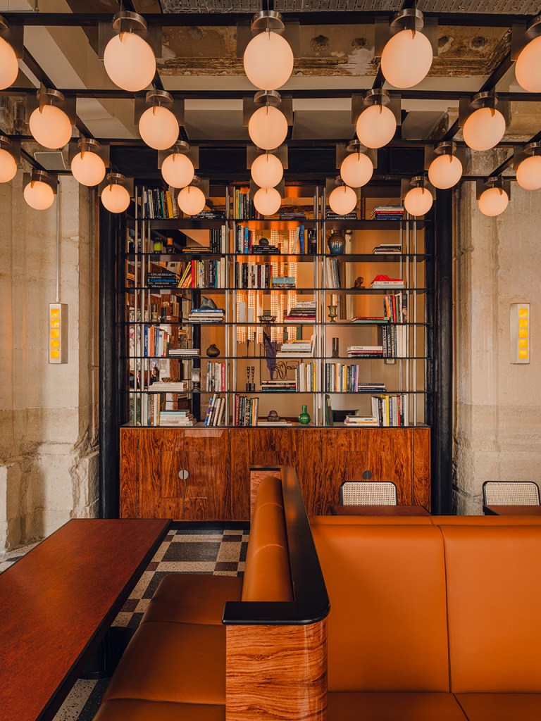 Modern hotel lobby with orange seating, wooden shelving unit filled with books, and pendant globe lighting.
