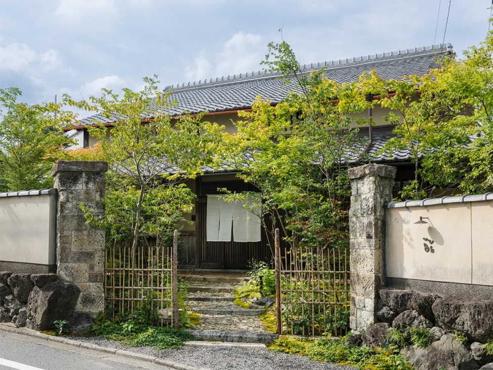 Traditional Japanese building with curved tile roof, stone pillars, metal gate, and lush green foliage in Kyoto.