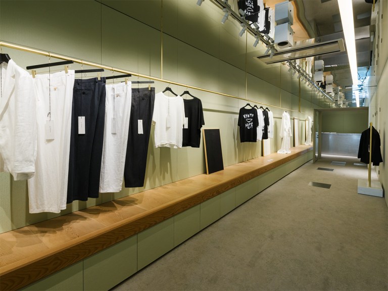 Minimalist retail space with sage green walls displaying clothing on brass rails and wooden counters.