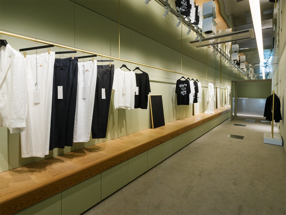 Minimalist retail space with sage green walls displaying clothing on brass rails and wooden counters.