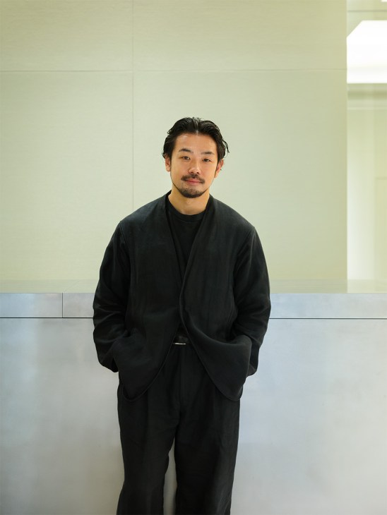 Masataka Hosoo, 12th-generation owner, wearing a black suit against a minimal geometric wall