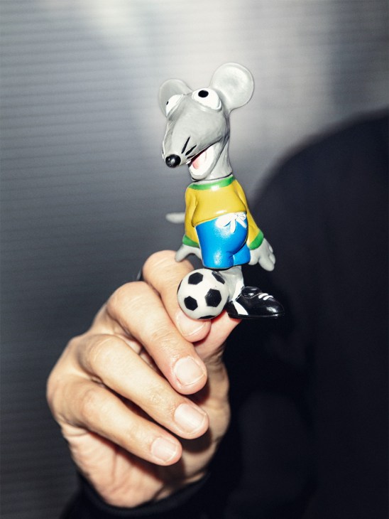 A hand holding a white mouse figurine wearing a yellow shirt with blue shorts and black shoes, with a soccer ball at its feet.