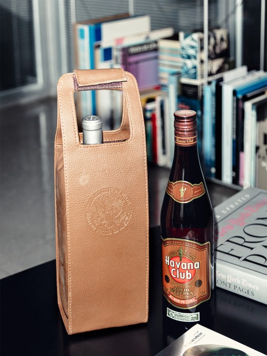 Havana Club rum bottle next to a tan leather wine carrier on a desk with books in background.
