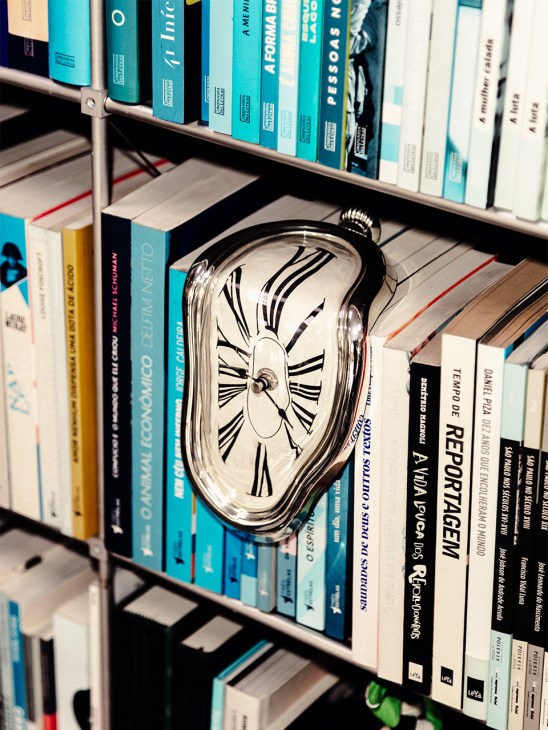 A melting clock with Roman numerals sits on a bookshelf among colorful books, referencing Salvador Dalí's 'The Persistence of Memory'.