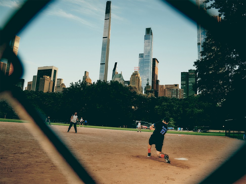 Baseball player batting at Heckscher Ballfields in Central Park with Manhattan skyline in background, framed through a chain-link fence.
