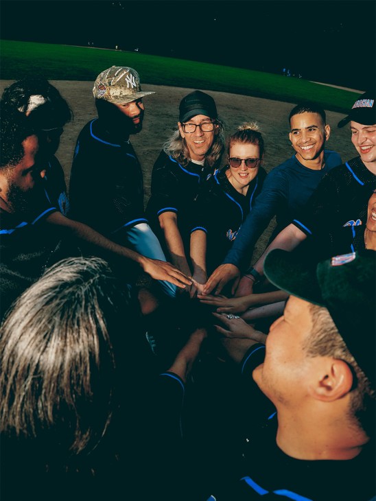 Museum and arts staff in blue uniforms huddle together with hands joined in a team circle at a night softball game.