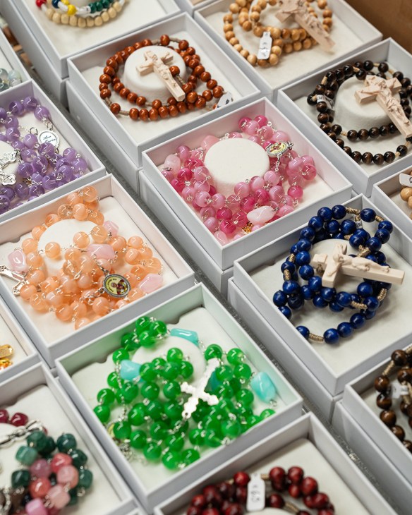 Display boxes of colorful beaded rosaries in various colors including pink, green, blue, brown, and orange with ivory cross charms at Savelli Religious shop.