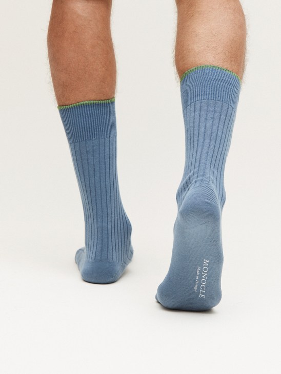 Man wearing blue cotton ribbed socks with green and yellow stripe detail and Monocle branding on the sole.