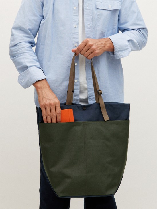 Person wearing a light blue shirt holding a navy and green canvas tote bag with tan handles and an orange item inside.