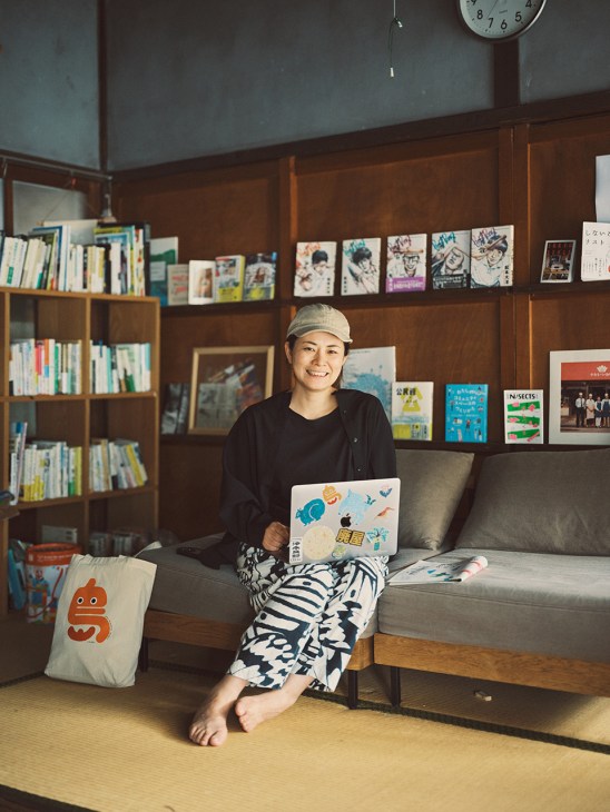 Atsuko Isamoto, editor in chief of Ritokei newspaper, sitting on a couch with a laptop surrounded by bookshelves and island-themed artwork.