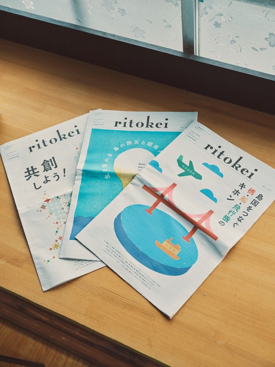 Three issues of Ritokei magazine displayed on wooden surface, featuring minimalist cover designs with illustrations of islands, aircraft, and water scenes in blue and red tones.