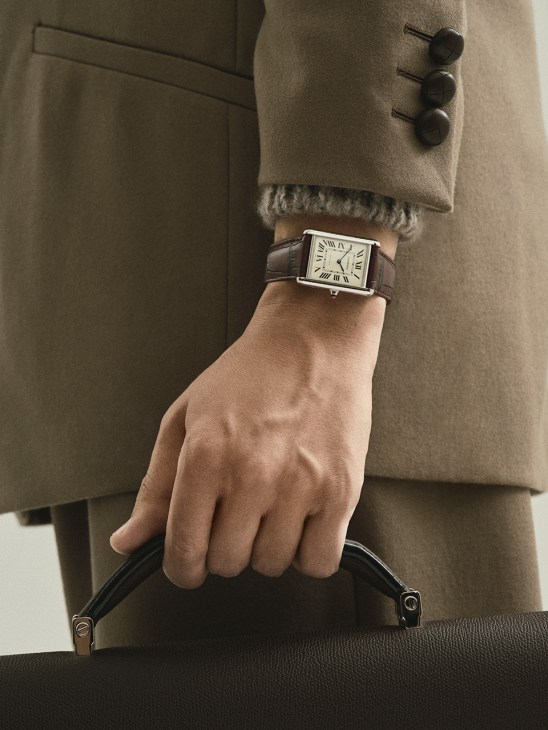 Wrist wearing Tank Louis Cartier watch with brown leather strap, beige wool suit and black handbag.