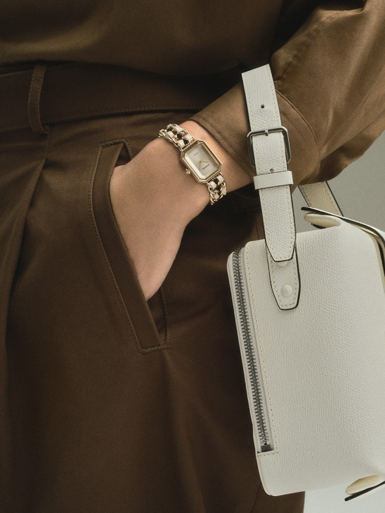 Person wearing a Chanel Première Iconic Chain watch with gold chain bracelet and silver rectangular face, styled with a brown Graphpaper shirt and white Valextra bag.