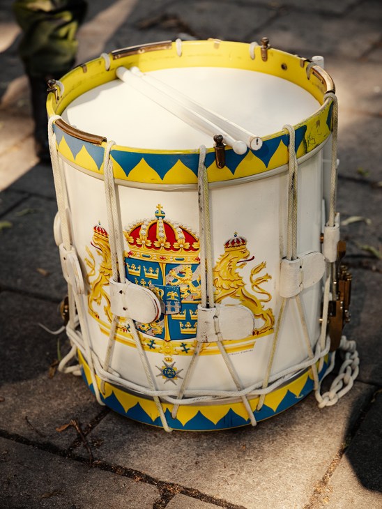 A decorative Swedish military drum with yellow and blue diamond pattern trim, white rope tuning, and royal Swedish coat of arms on the white shell.