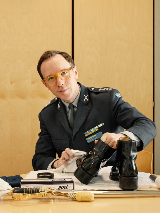 Military officer in black uniform with yellow glasses examining black military boots at a table