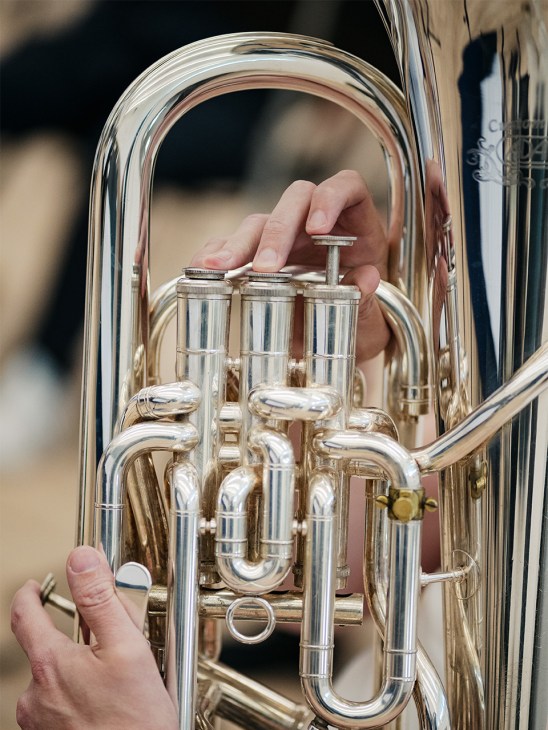 Hands holding a polished brass tuba valve mechanism up close during a performance.