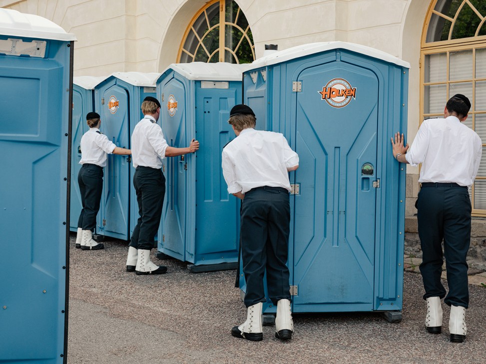 Swedish Army Band musicians in white shirts and dark trousers using blue portable toilets labeled Houken.