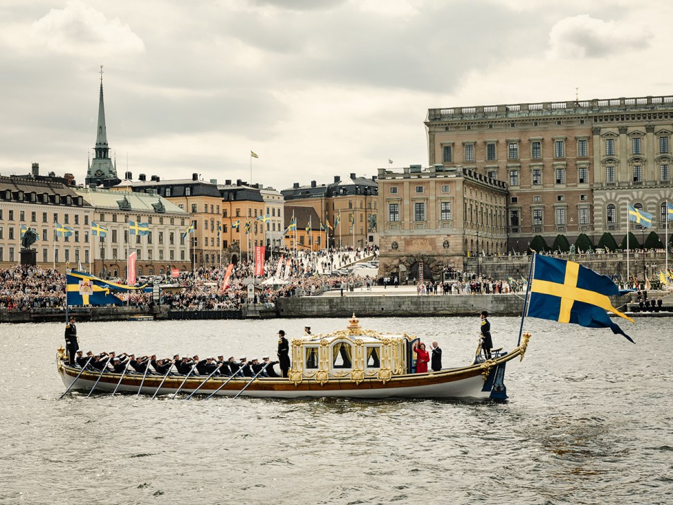 Ornate royal barge with gold canopy on Stockholm harbor water, Swedish flags, crowds and historic buildings in background.