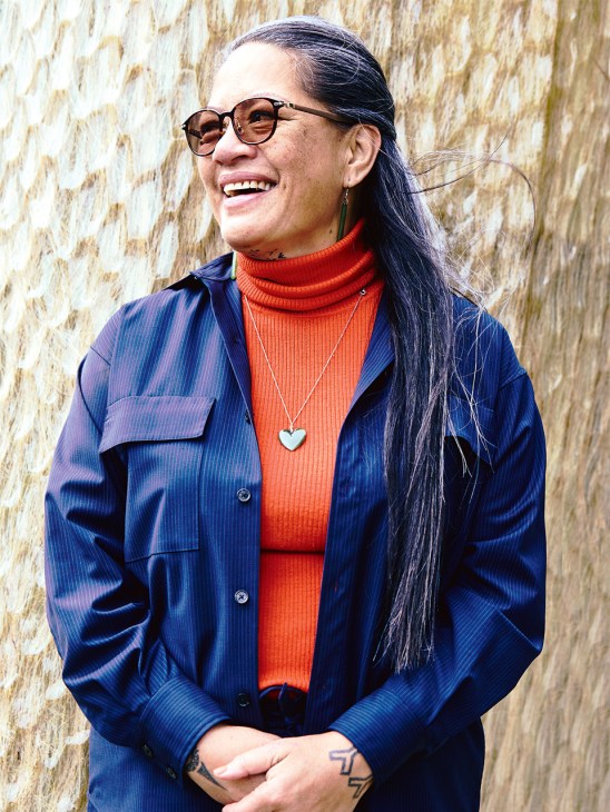 Lucy Tukua, Mana Whenua advocate, smiling against a textured brick wall wearing glasses, red turtleneck and blue jacket.