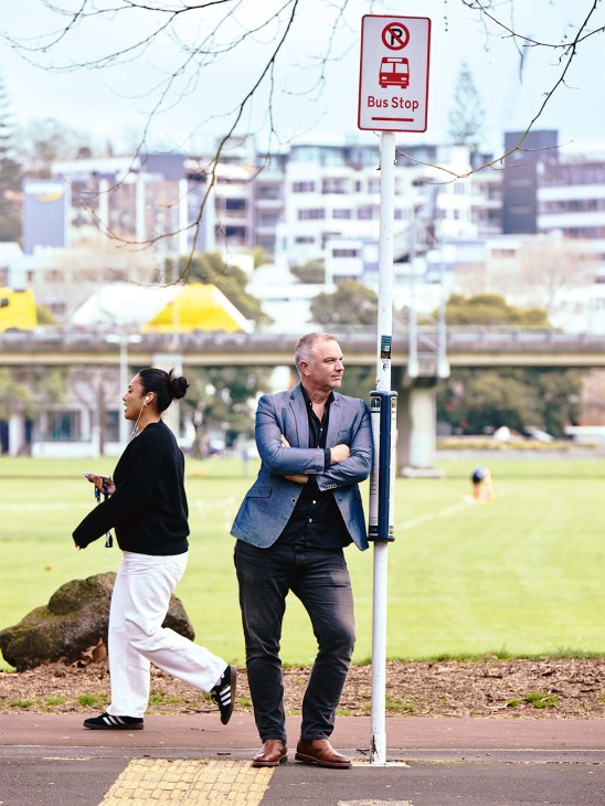 Man in blue blazer standing at a bus stop sign near a waterfront park and apartment buildings.