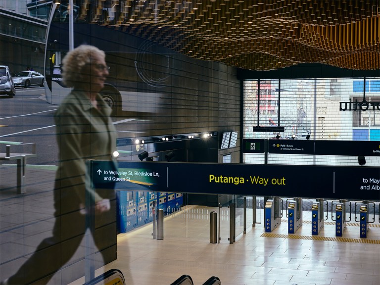 Te Waihorotiu station interior with yellow signage, ticket gates, and a blurred figure walking past glass panels.