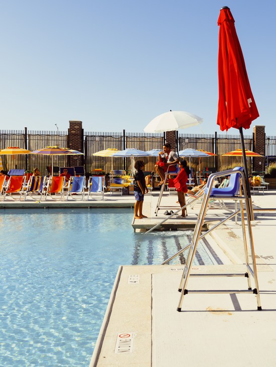 Guests lounging by a U-shaped swimming pool at Rockaway Ocean Club with colorful striped deck chairs and red umbrellas.