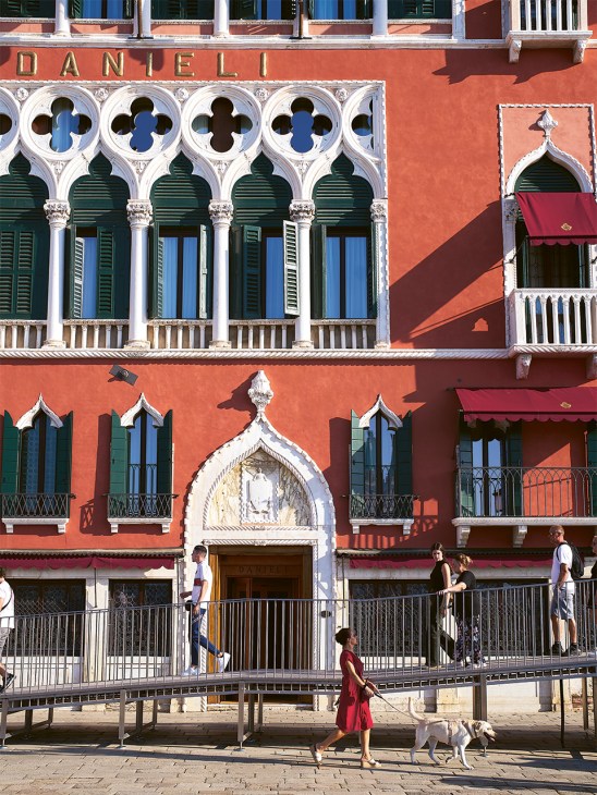 The Danieli hotel's ornate red Gothic façade with white stone arches, green shutters, and people walking along the waterfront promenade below.