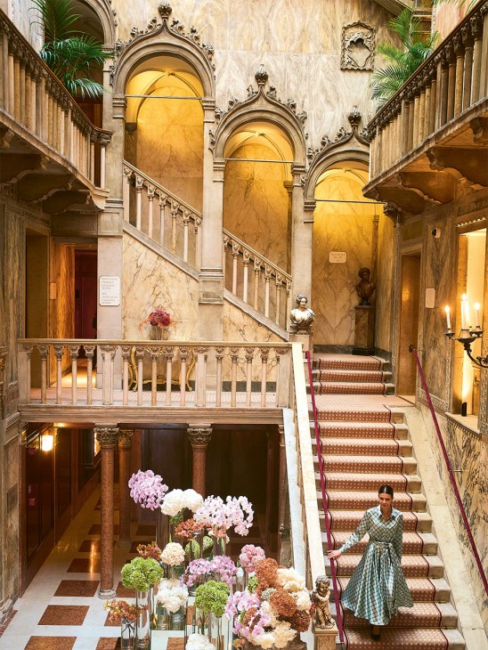Ornate marble staircase with patterned carpet and decorative railings in the Danieli hotel's central atrium, with classical columns and arched alcoves.