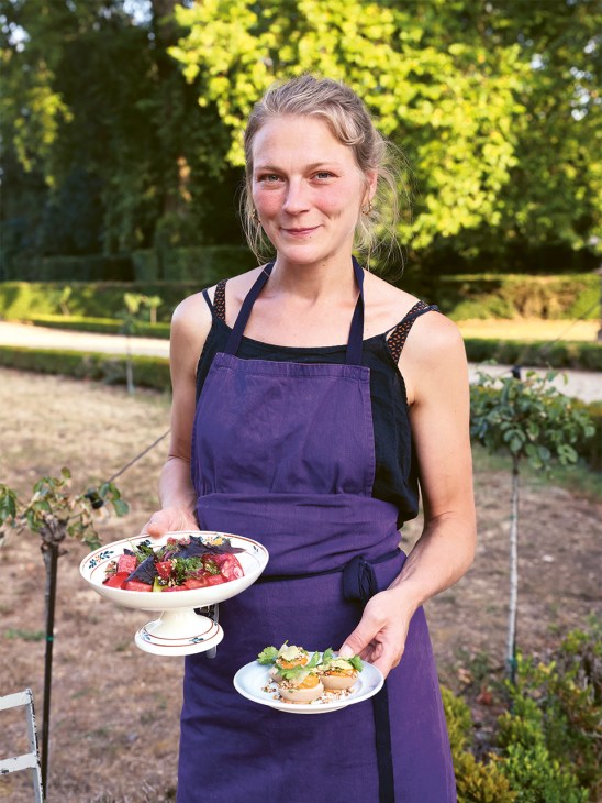 Sommelier Ioulia Gourieva holds two plated dishes in a garden setting, wearing a purple apron.