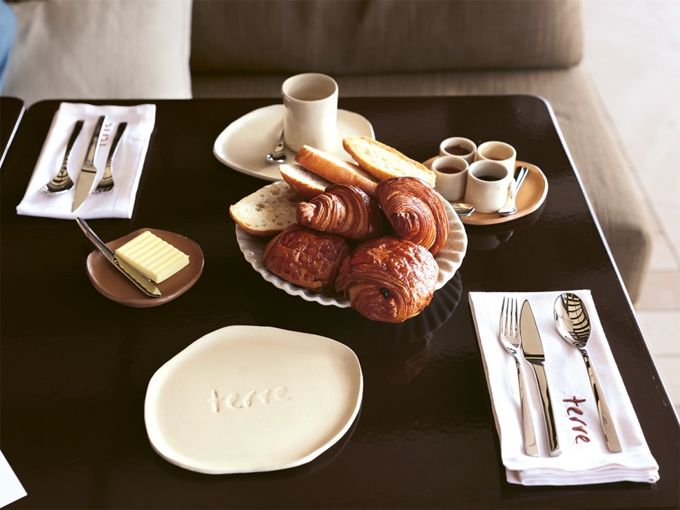 Breakfast table setting at Maison Le Chevreuil with croissants, pastries, bread, butter, coffee cups, and place settings on a black table.