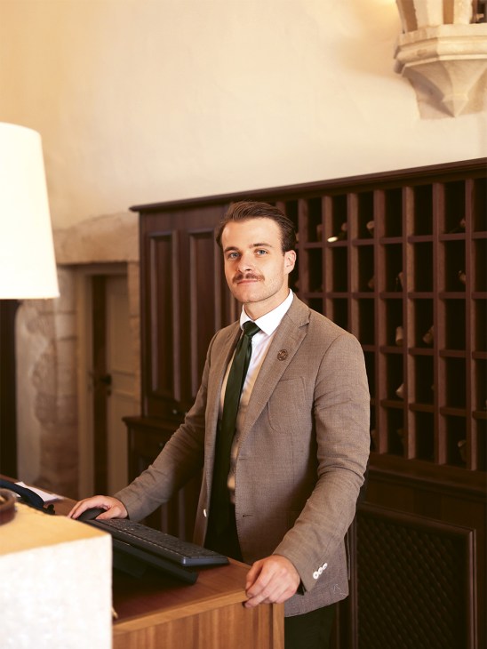 Man in beige blazer and dark tie standing at reception desk of Les Sources de Vougeot hotel.