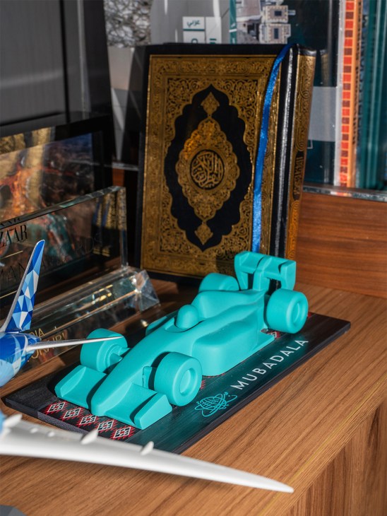 Turquoise Formula 1 race car model on dark base with Mubadala branding, displayed next to decorative Quran and magazines on wooden desk.