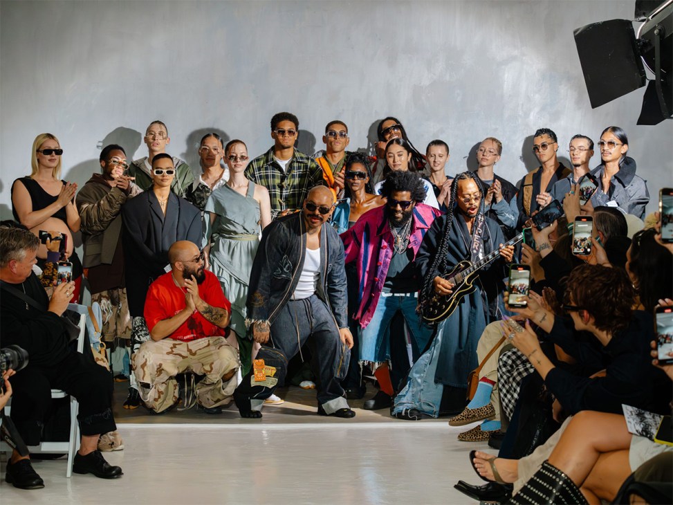 A diverse group of people stands and sits against a white wall at Atelier Reserve during Amsterdam Fashion Week, with audience members photographing the event.