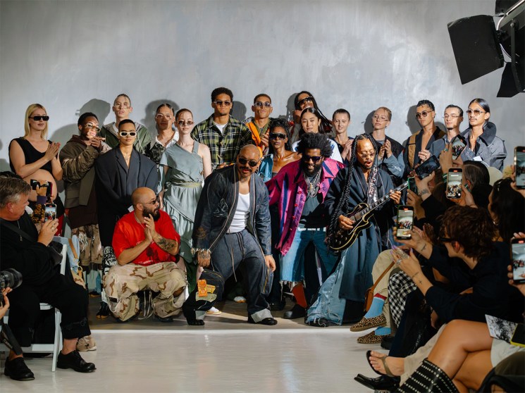 A diverse group of people stands and sits against a white wall at Atelier Reserve during Amsterdam Fashion Week, with audience members photographing the event.