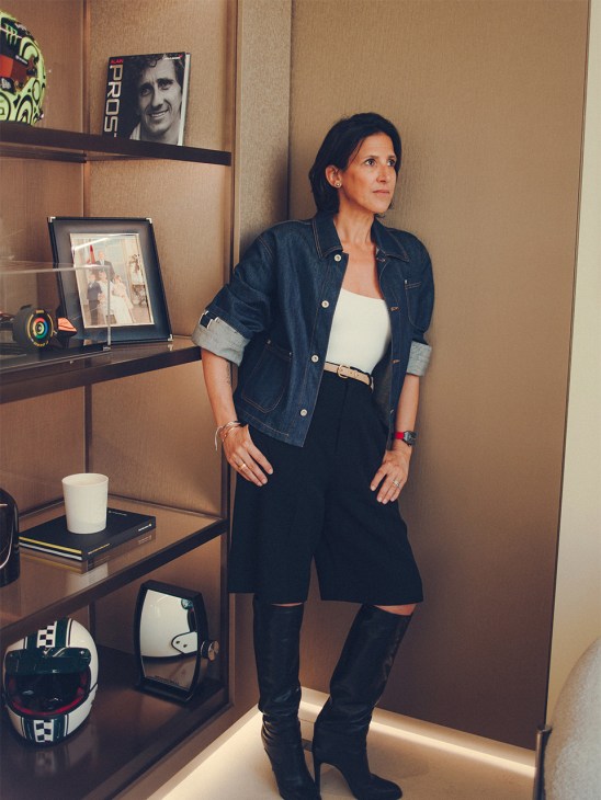 Woman in denim jacket and black ripped pants standing in front of shelving with watches and memorabilia in an office setting.