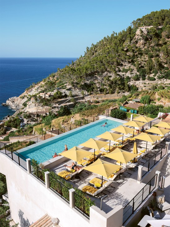 Gran Hotel Margalida's turquoise pool with yellow umbrellas and loungers overlooking the Mediterranean Sea and cliffside terraces of Banyalbufar, Mallorca.