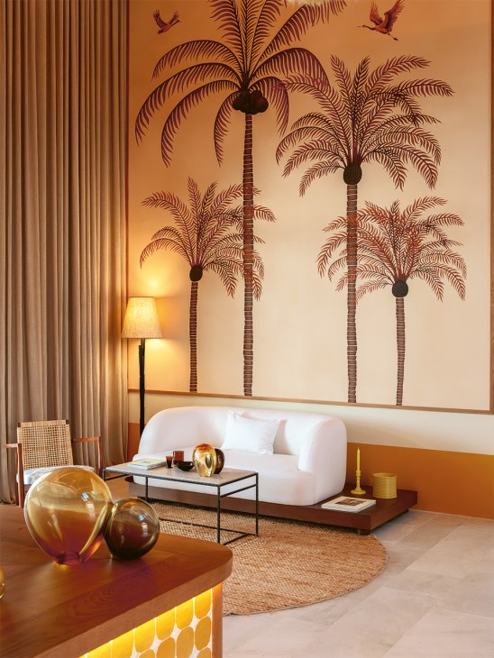 Mediterranean hotel suite with white curved sofa, palm tree wall mural, and ambient lighting designed by Virginia Nieto.