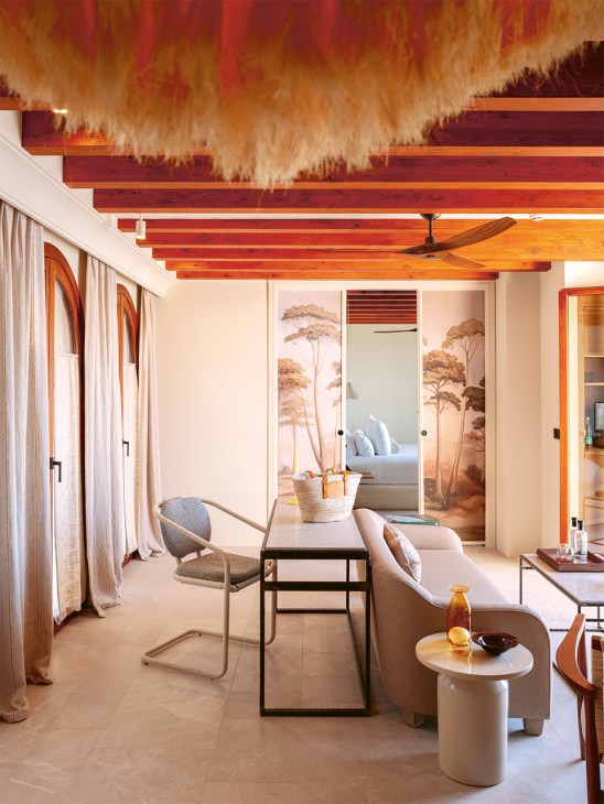 Hotel suite with cream-colored furniture, wooden beamed ceiling, shaggy yellow light fixture, and palm tree wall murals.