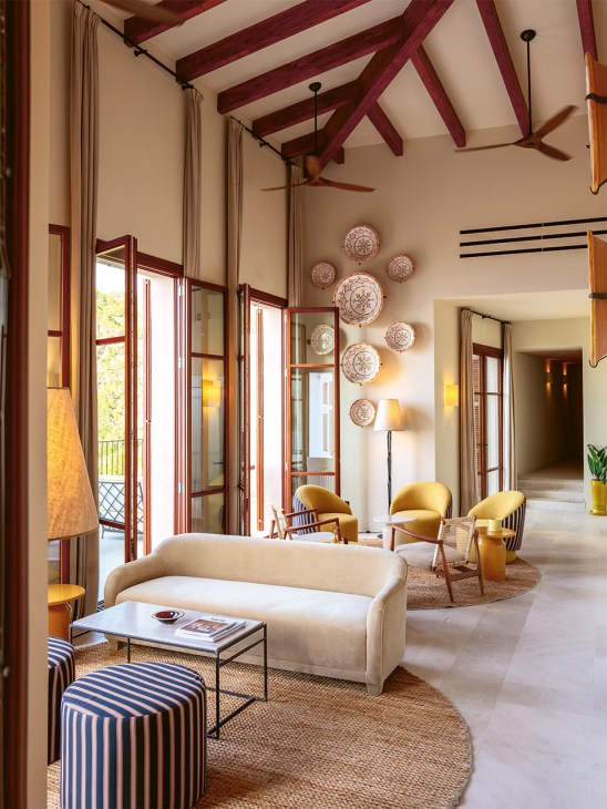 Salon at Gran Hotel Margalida with cream sofa, striped pouf, decorative plates on wall, and yellow chairs visible in background.